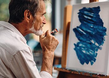 Memory Care Services. Mature painter holds a paintbrush while looking at his artwork.